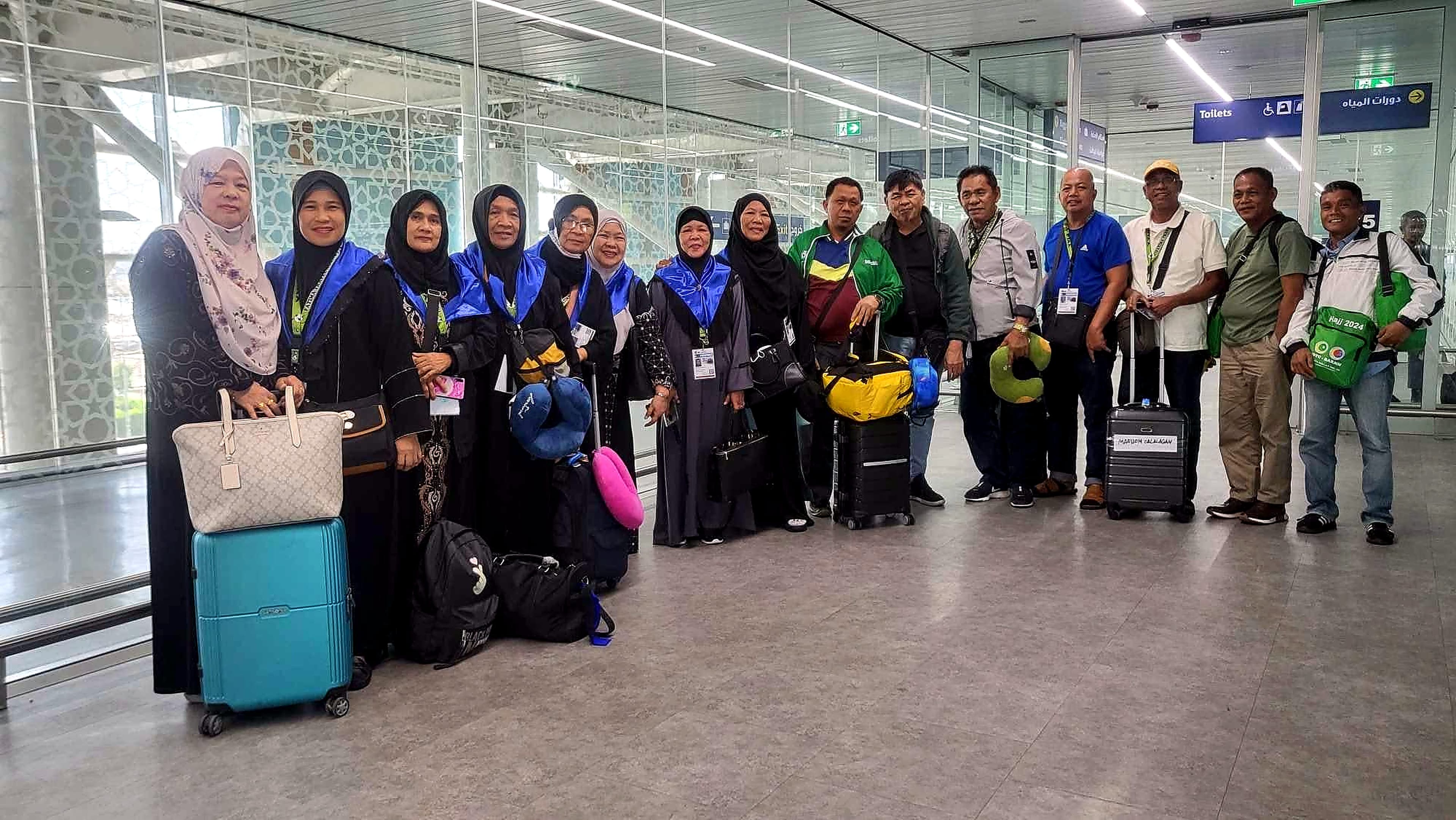 A group of pilgrims with lanyards and luggage standing together inside an airport terminal.