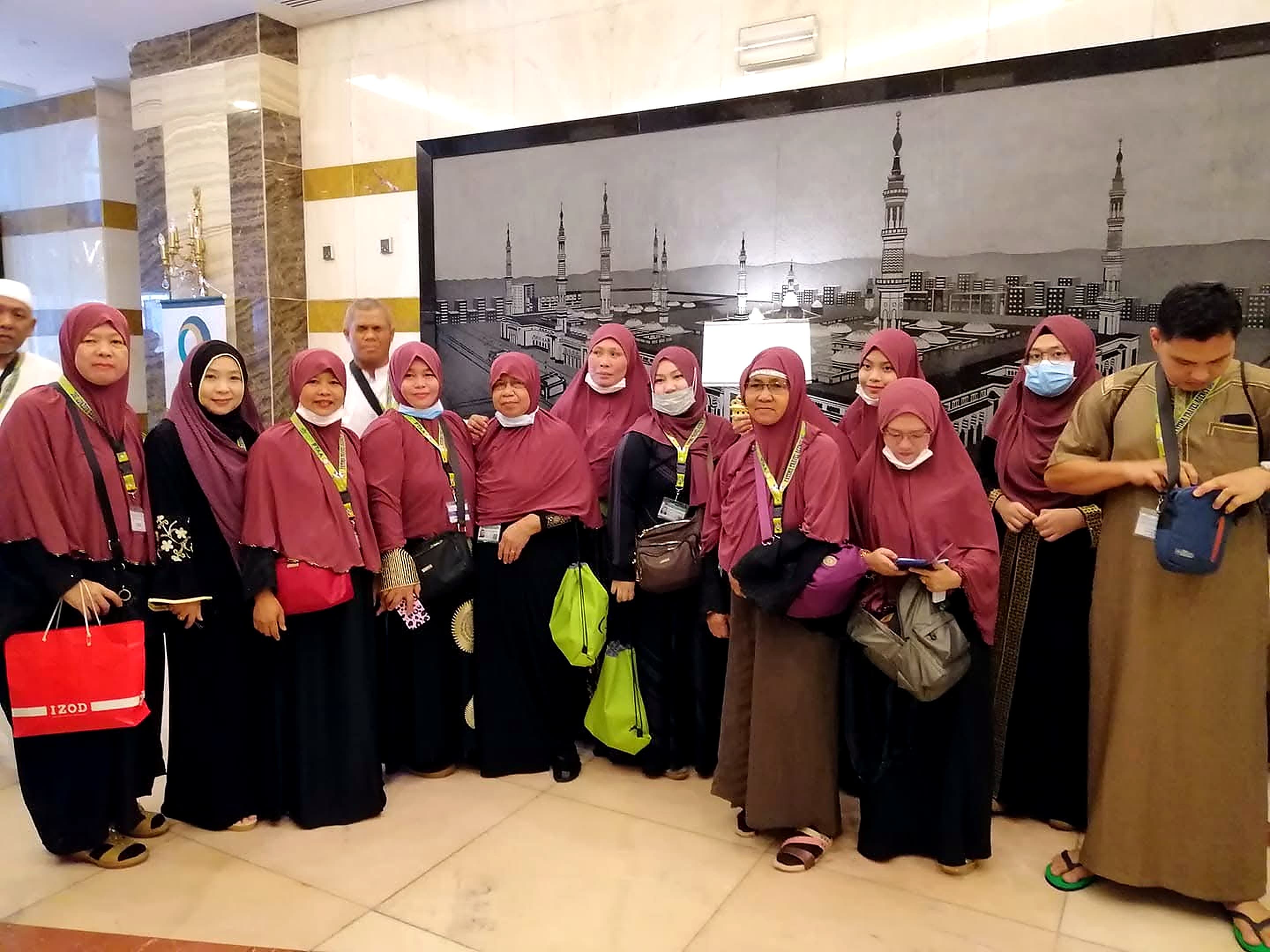 A large group of women pilgrims in matching maroon headscarves gathered before a mural of a mosque with tall minarets.