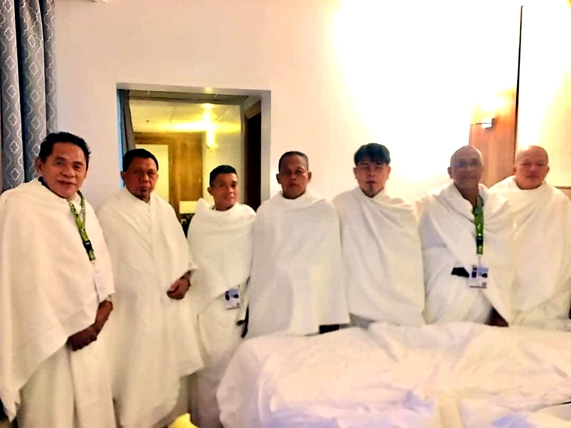 Seven men in white ihram garments standing side by side in a hotel room.