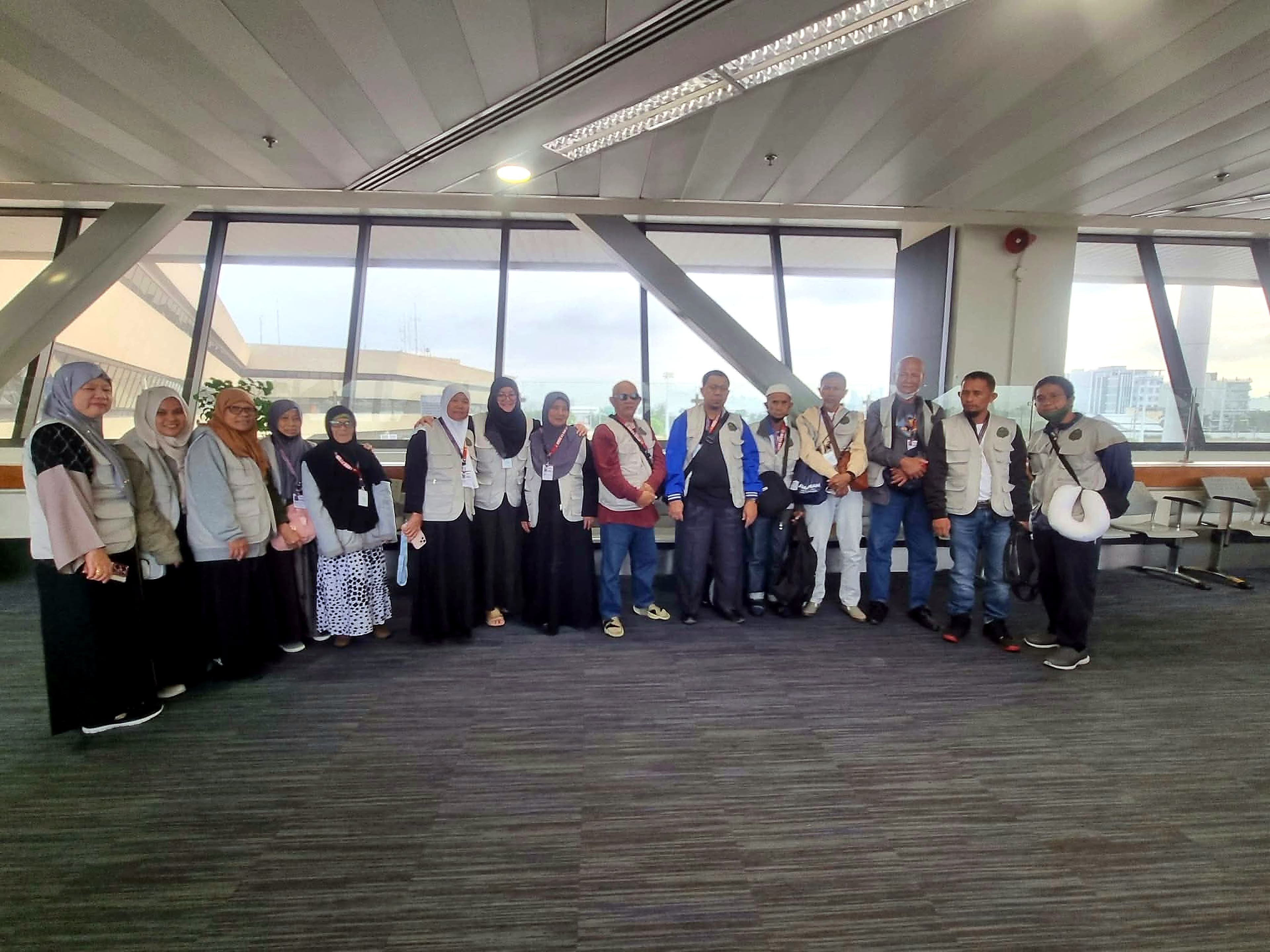 Pilgrims in matching vests lined up together beside large windows in an airport terminal.