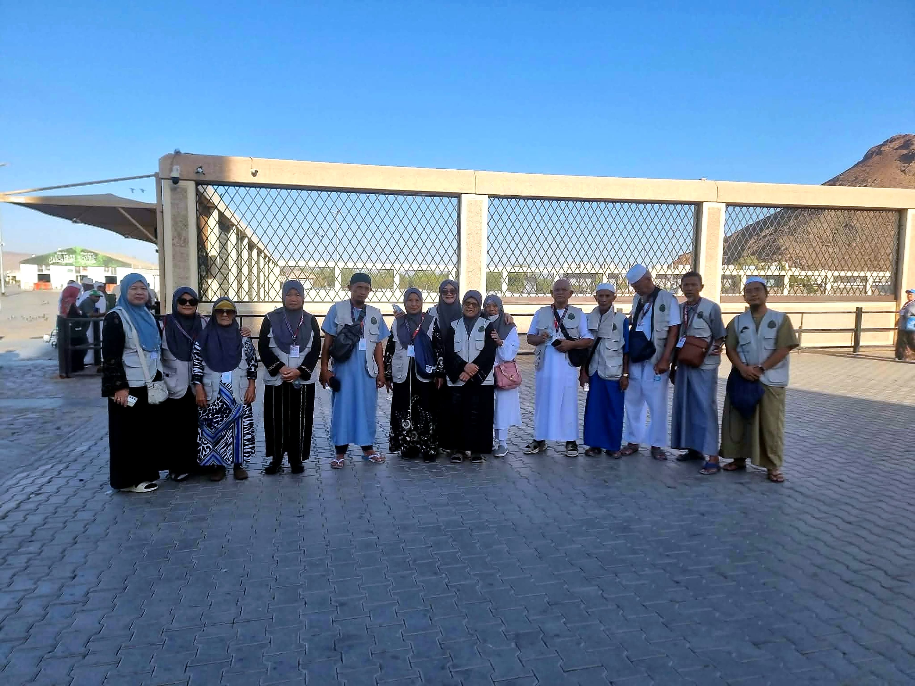A group of pilgrims in matching vests standing in a row on a paved plaza under a clear blue sky.