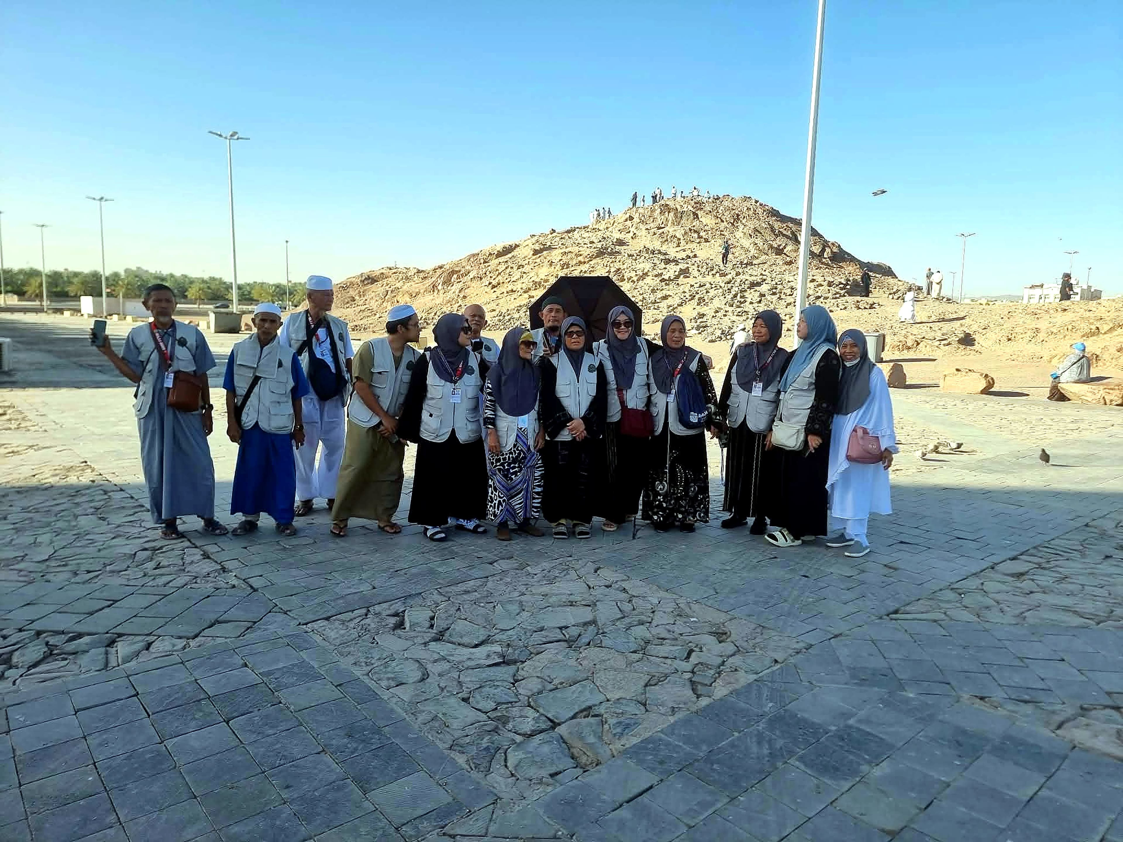 Pilgrims in matching vests gathered on a stone plaza, with a rocky hill and other visitors behind them.